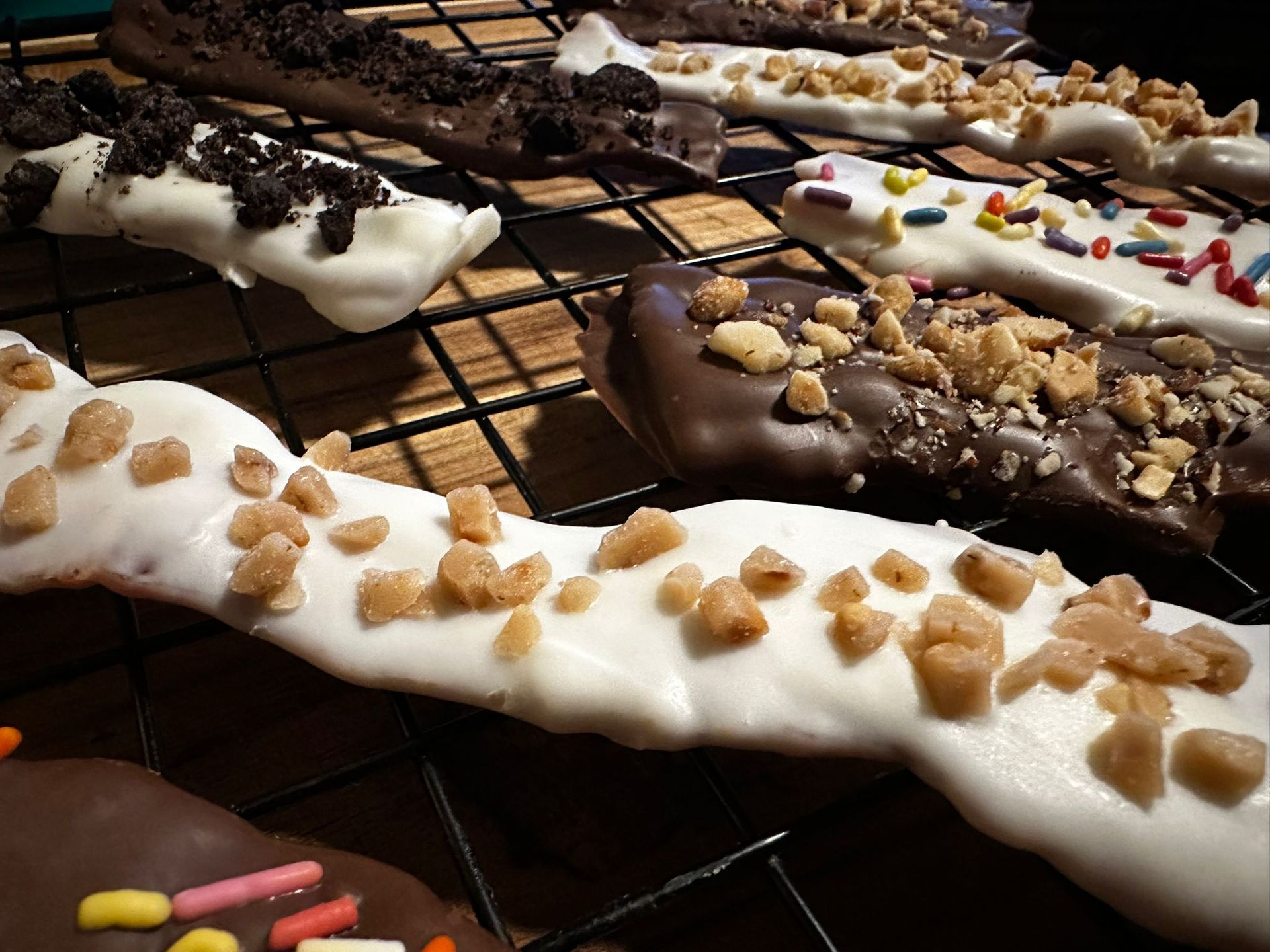 Chocolate-covered pretzel rods, some with sprinkles, crushed cookies, and toffee bits, cooling on a wire rack.