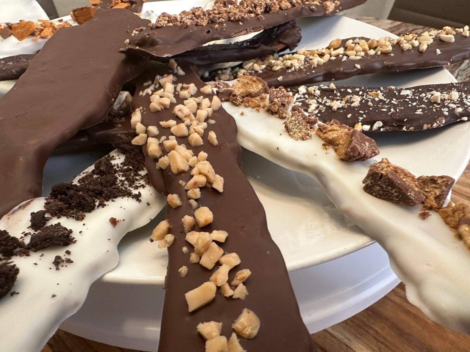 Chocolate-covered pretzel rods, some with toppings like toffee, nuts, and cookie crumbles, displayed on a white plate.