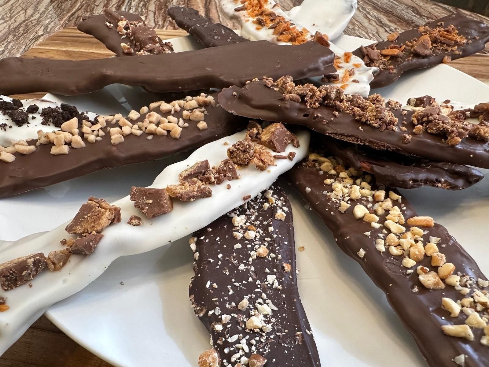 Chocolate-covered pretzel sticks, dark and white chocolate, topped with crushed candies and nuts, arranged on a white plate.