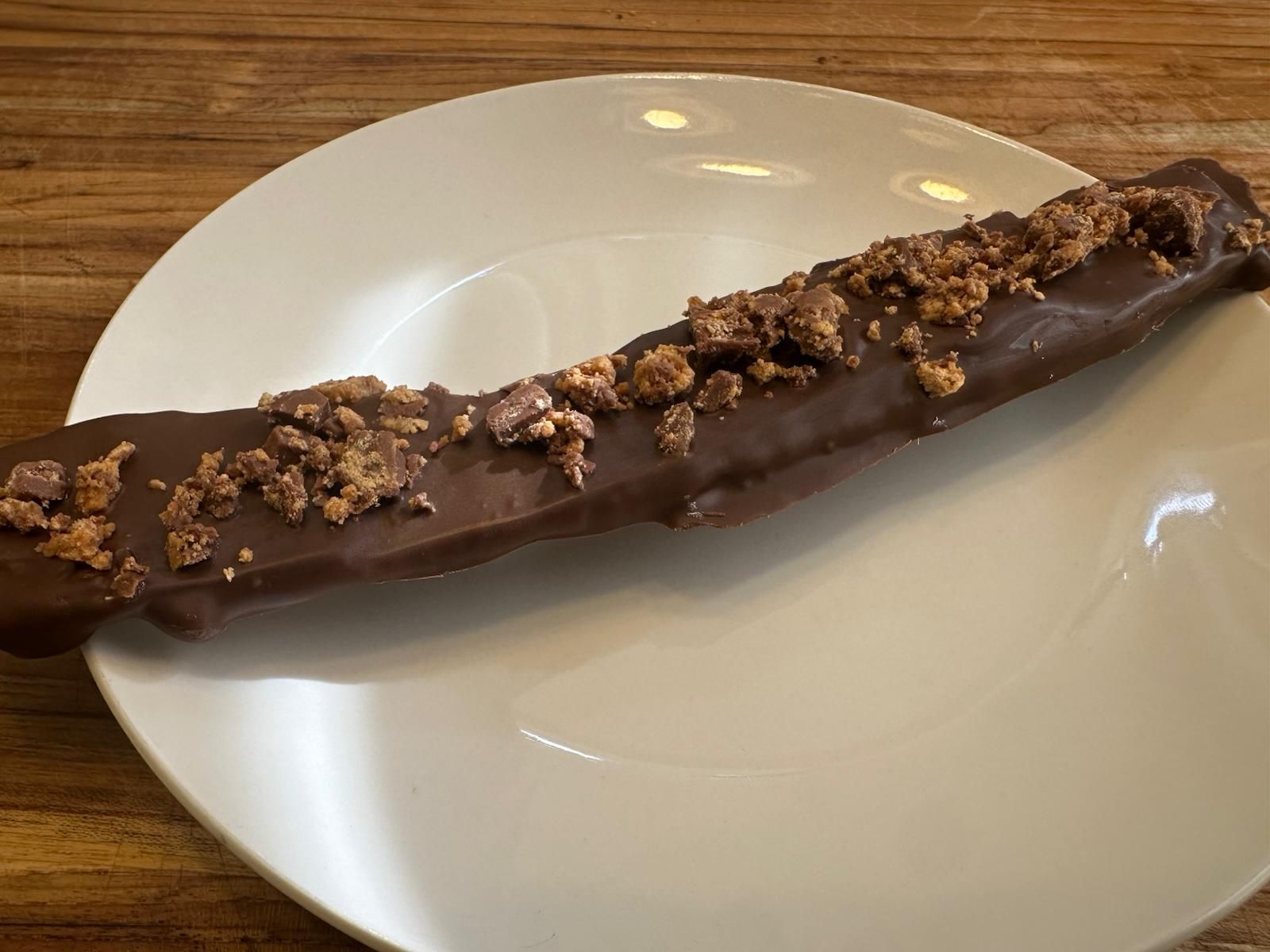 Chocolate-covered frozen banana on a white plate, topped with crushed chocolate pieces.