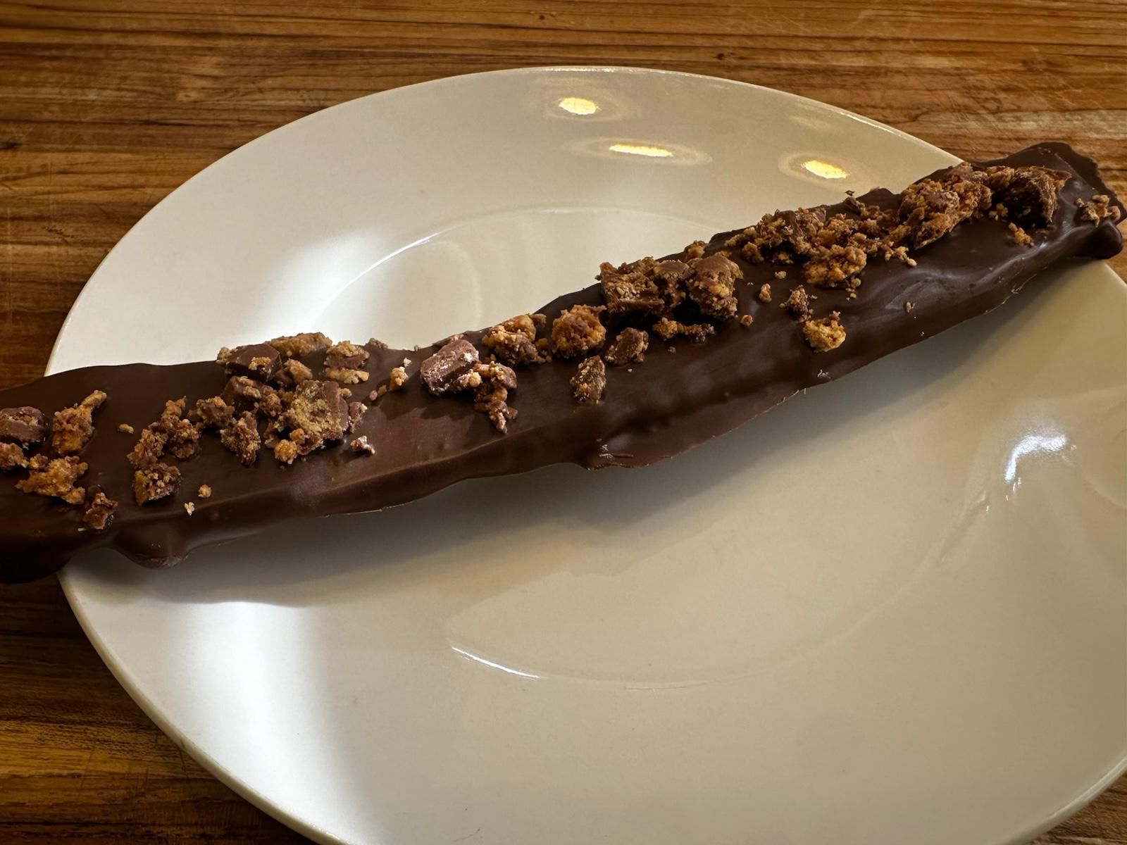 Chocolate-covered banana on a white plate, topped with crumbled pieces, set on a wooden table.