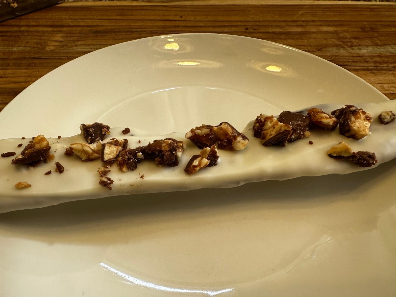 White-frosted éclair topped with chopped nuts and chocolate, resting on a white plate.