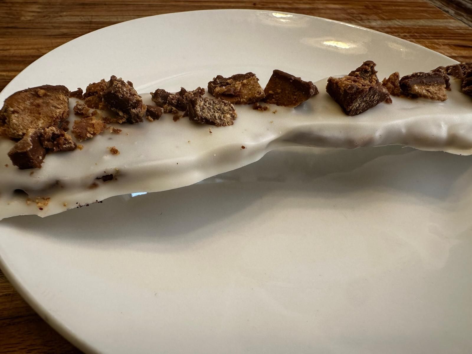 A white frosted pretzel stick topped with crumbled chocolate candy on a white plate.