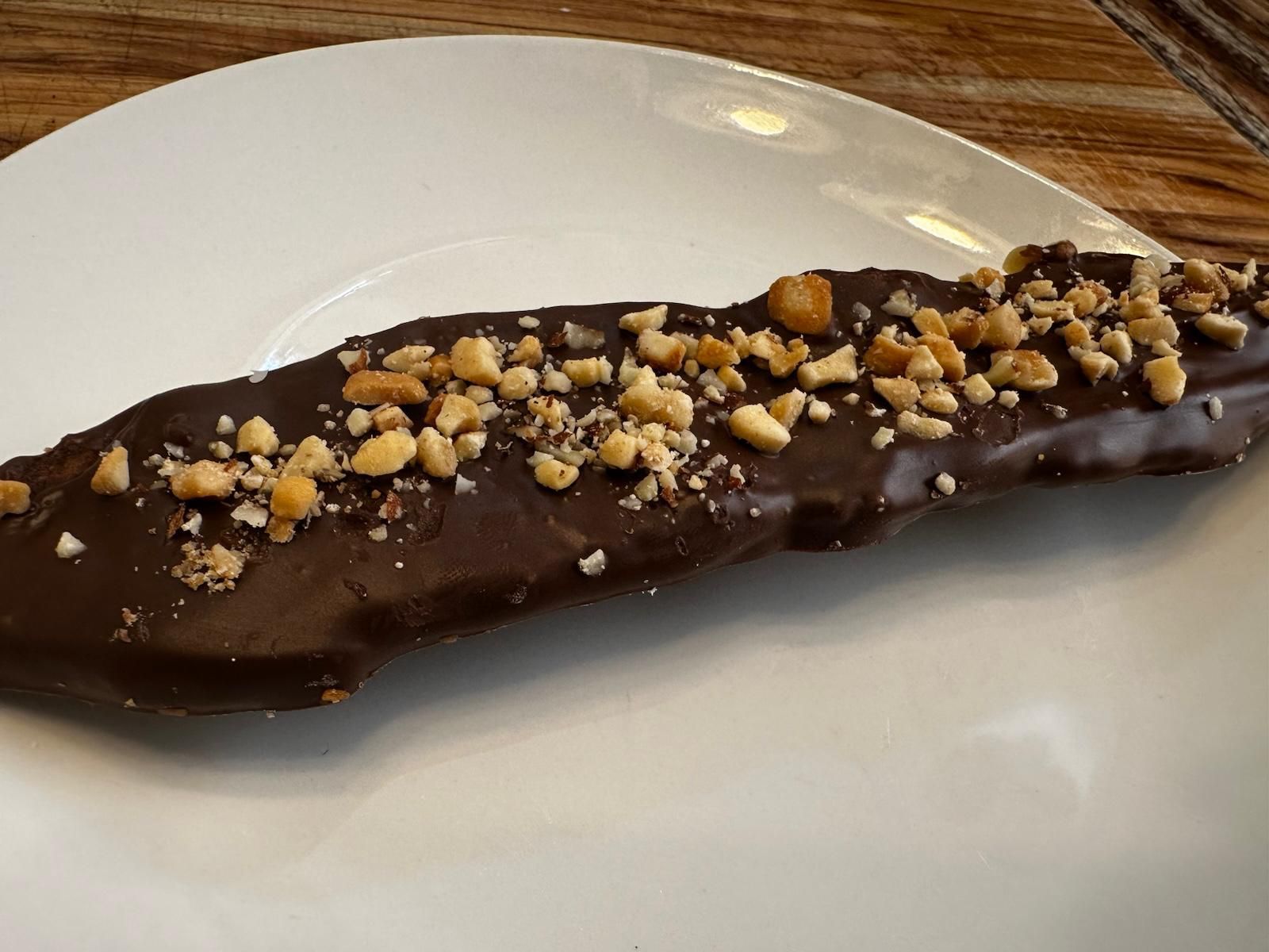 Chocolate-covered frozen banana topped with chopped peanuts on a white plate.