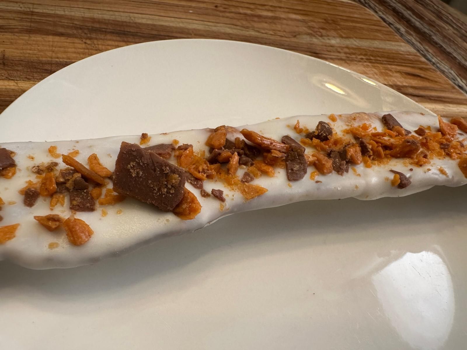 White-frosted pretzel rod topped with crushed toffee and chocolate pieces on a white plate.