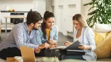 Couple consults with a professional; paperwork, laptop on table, bright interior.