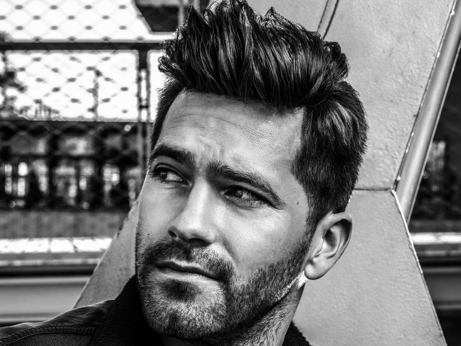 Man with styled hair and beard, looking off to the side, outside near a structure.