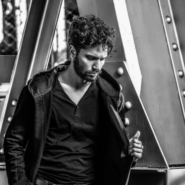 A man with a beard is standing in front of a metal structure in a black and white photo.