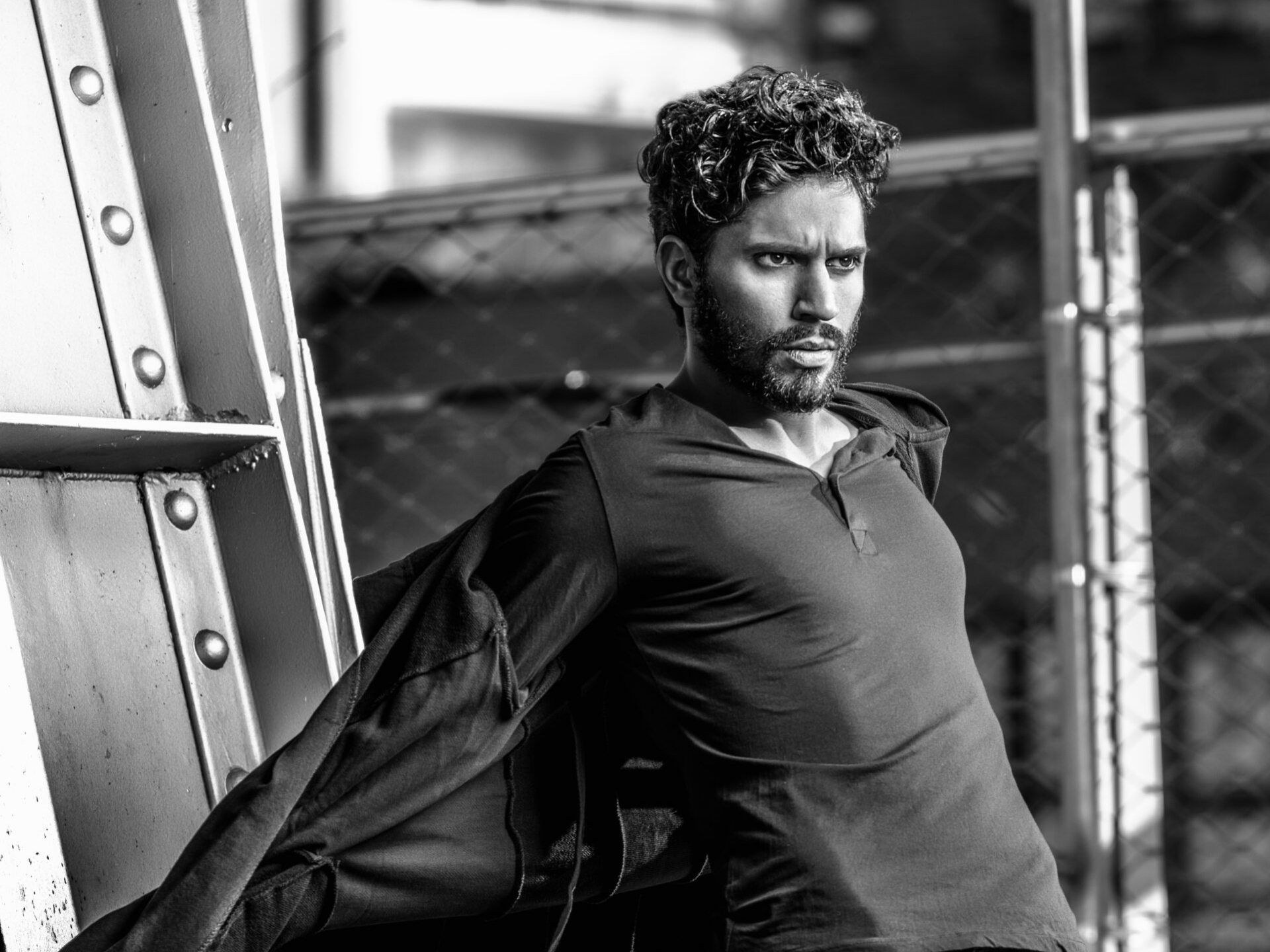Man leaning against metal structure, wearing dark shirt, looking off to the side with serious expression. Black and white.