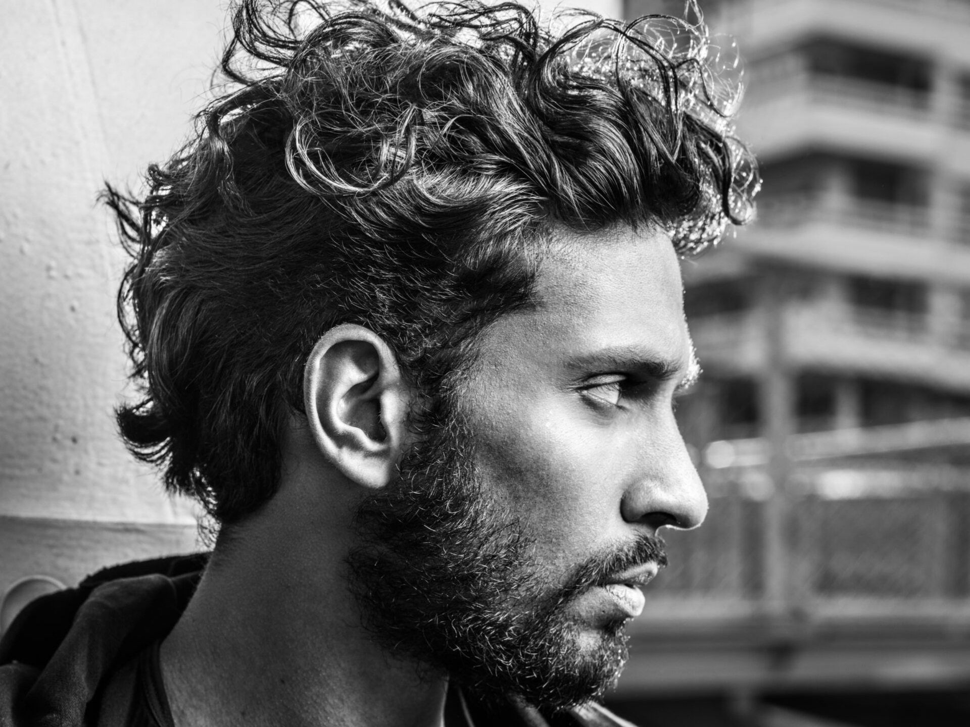 Man with curly hair and beard, looking away in profile, leaning against a wall in an urban setting.