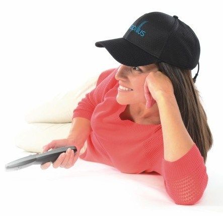 A woman wearing a black baseball cap is laying on a couch holding a remote control