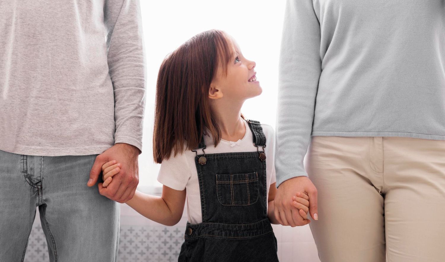 Girl smiles, holding hands with parents. They stand indoors, near a window.