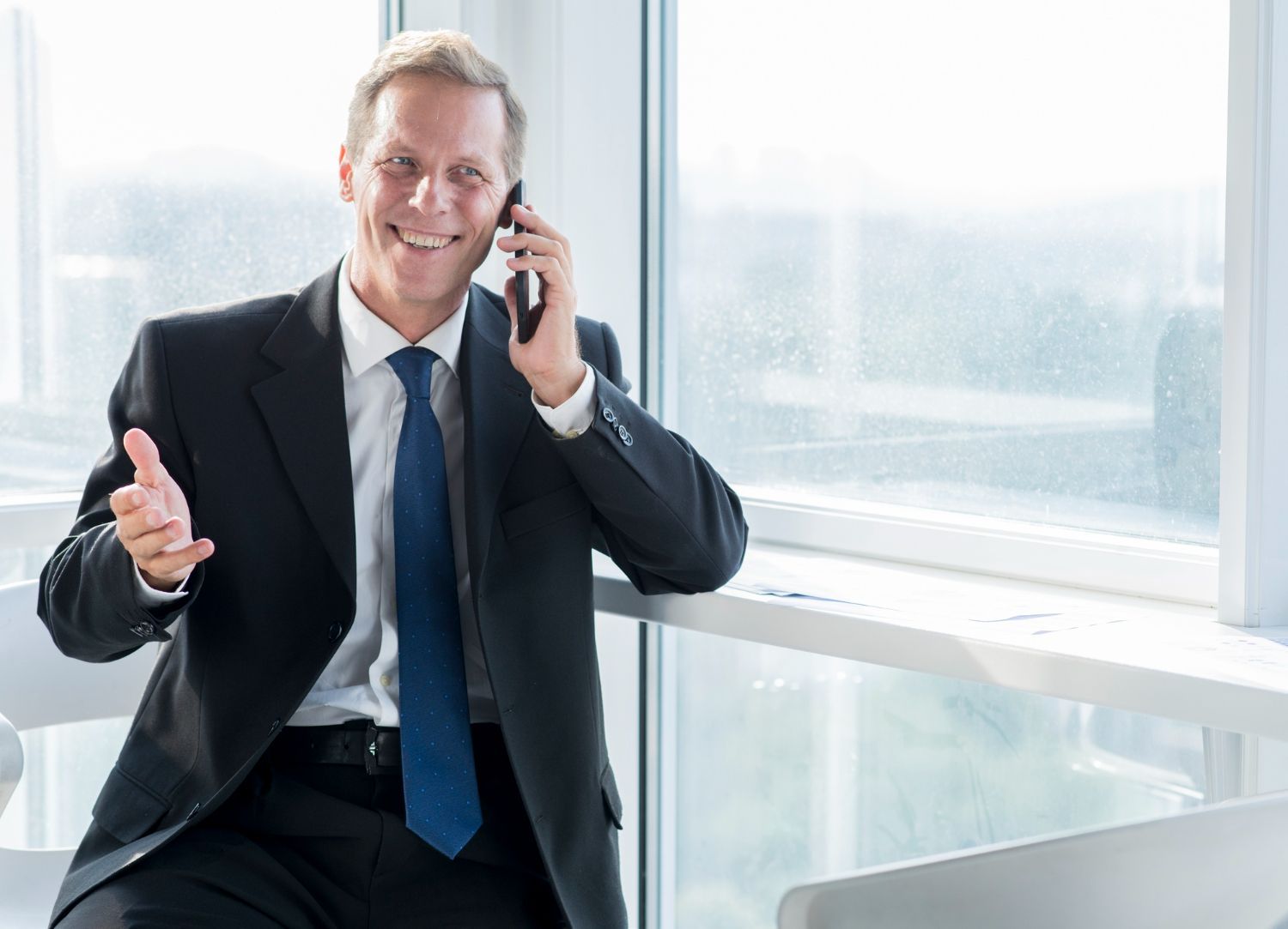 Smiling businessman in suit talking on phone near a window, gesturing with his hand.