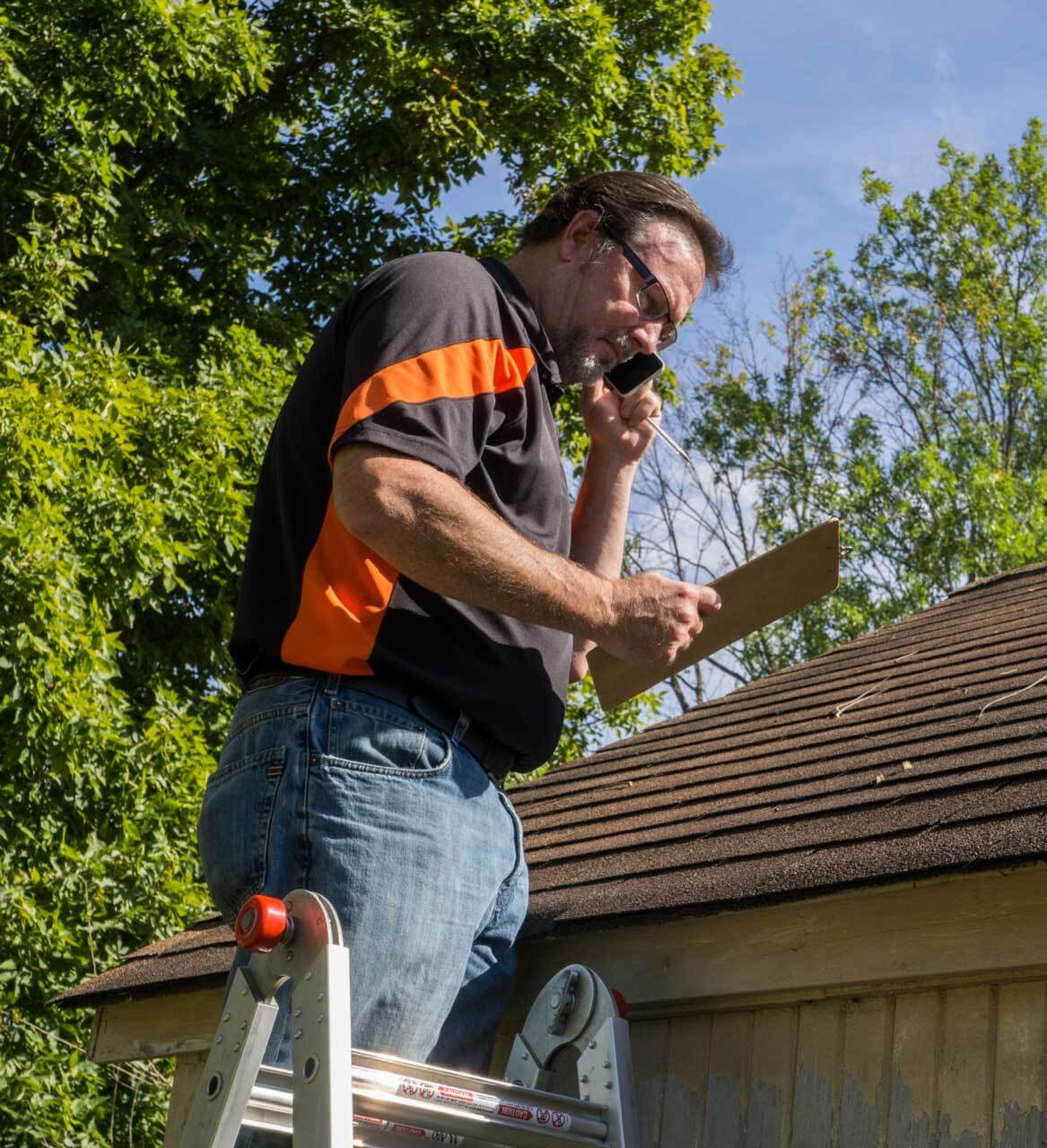 Contractor-on-ladder-with-cell-phone-figuring-out-hail-damage-repair-costs-for-customer