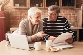 Happy-Senior-Couple-Planning-Family-Budget-Together-With-Laptop-And-Papers