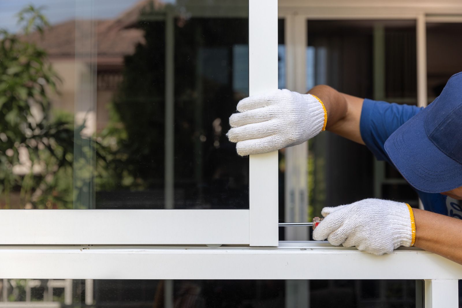 A worker wearing white work gloves uses a tool to install a white sliding glass door frame.