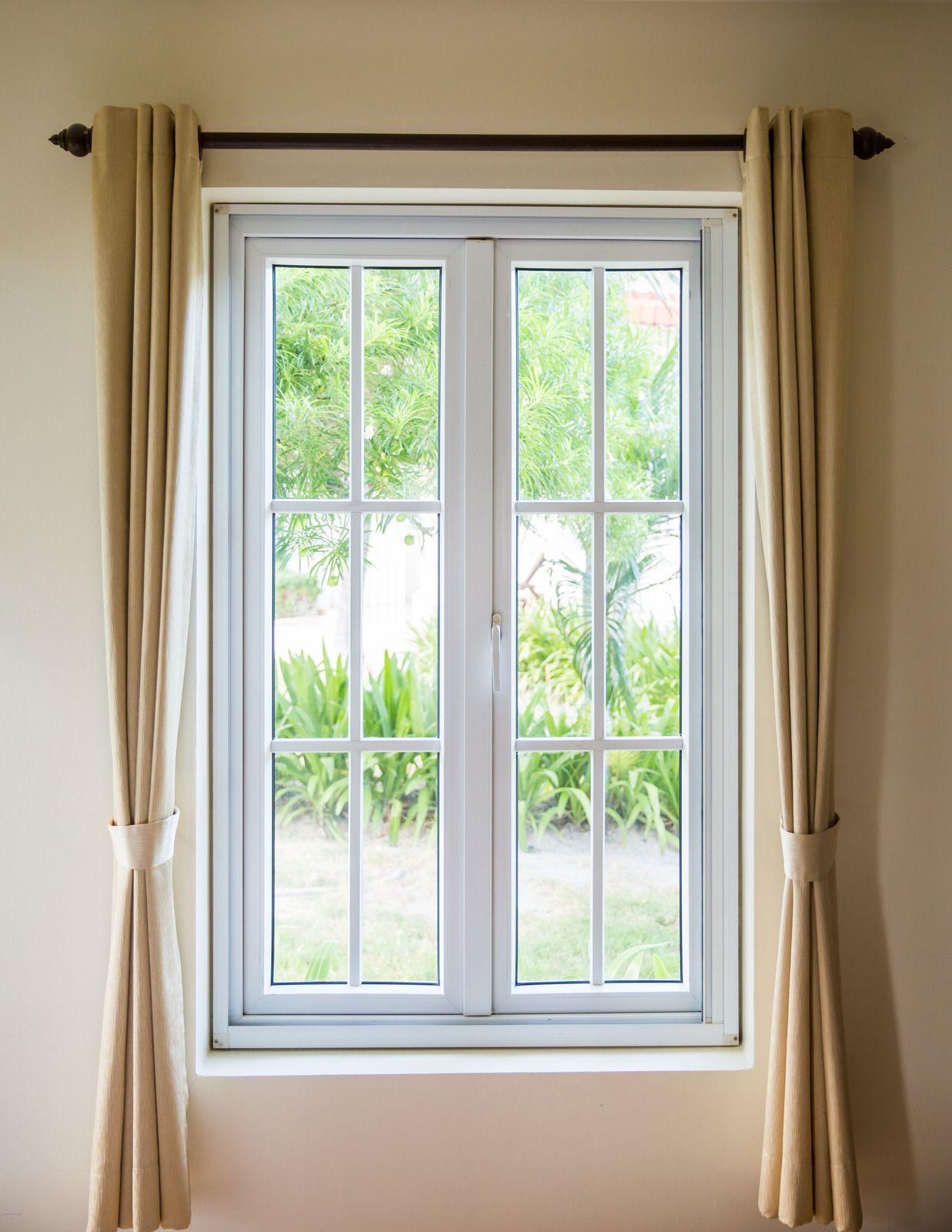 A white window with a grid pane design, framed by light beige curtains on a dark brown rod against a neutral wall.