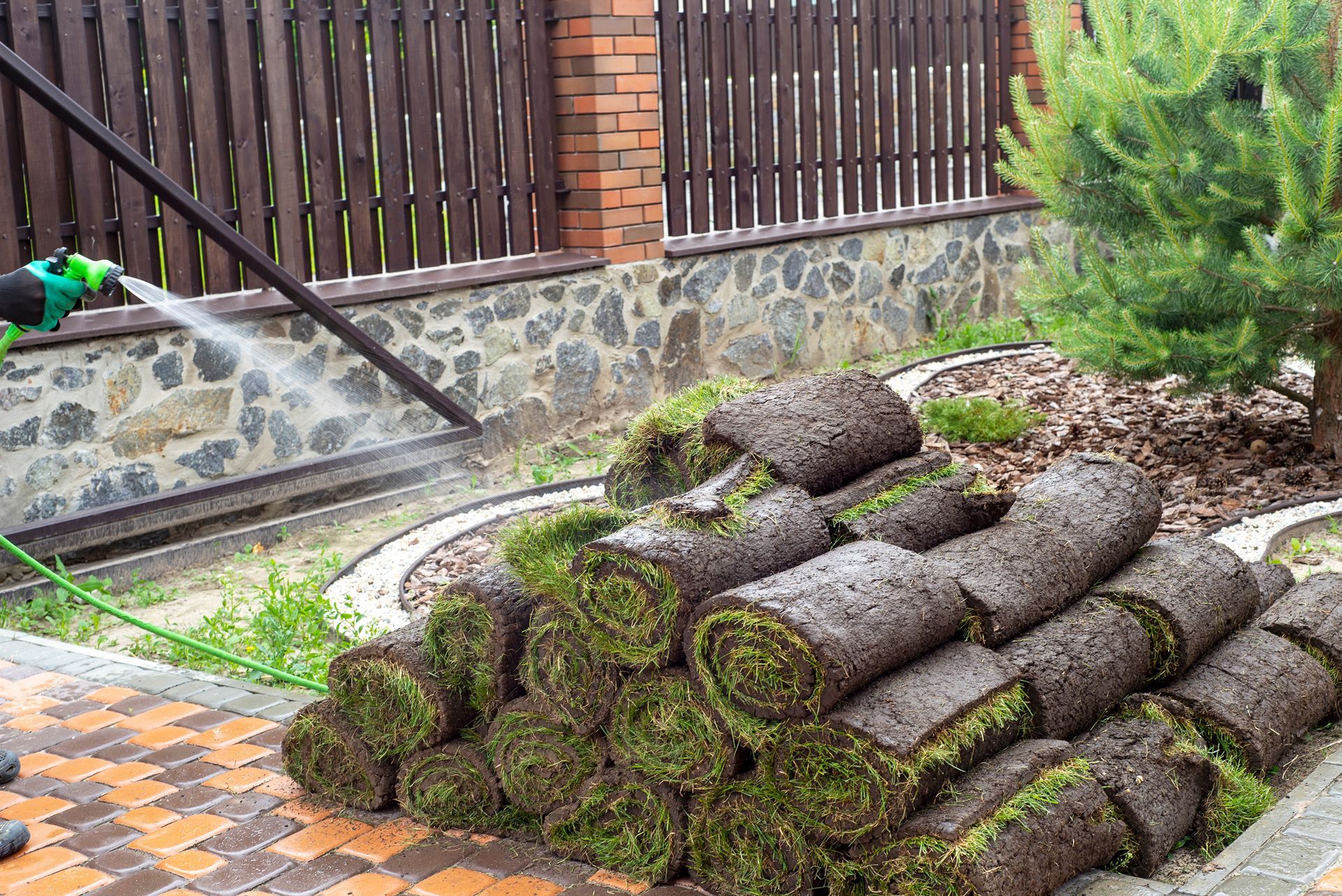 A hand uses a green spray nozzle to water a large, stacked pile of grass sod rolls beside a paved walkway and stone fence.