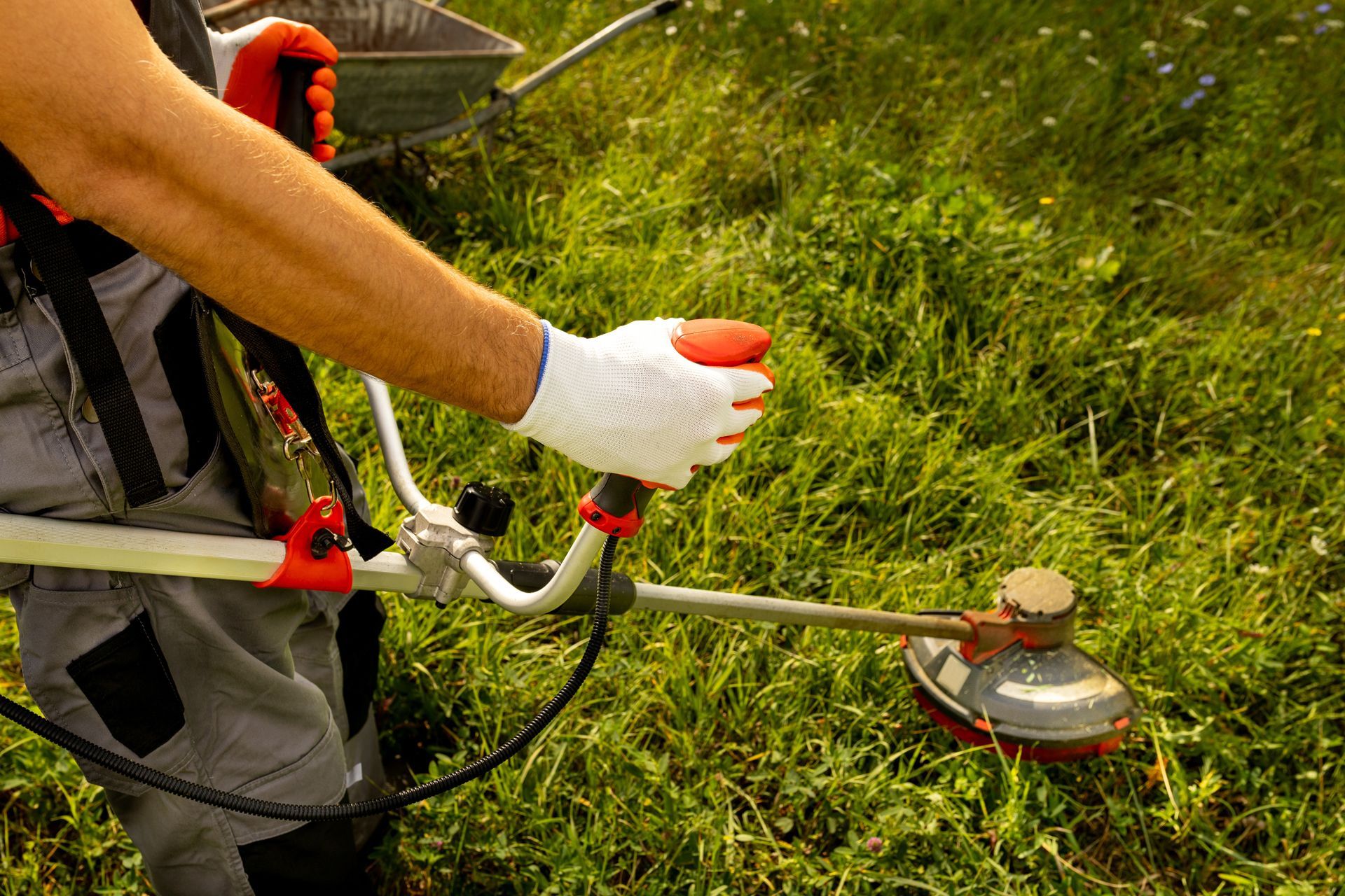 A person in work clothes and gloves uses a string trimmer to cut grass in a field.