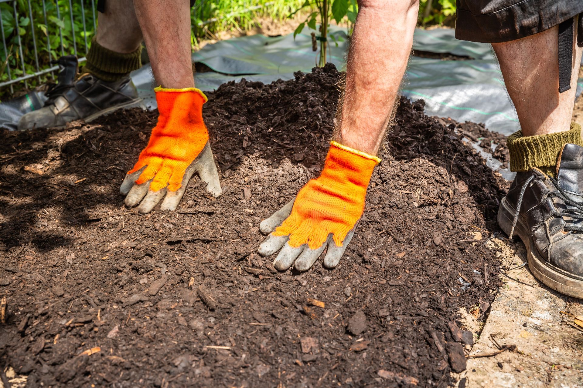 Person in work boots and bright orange gloves spreading dark soil on the ground in an outdoor garden.