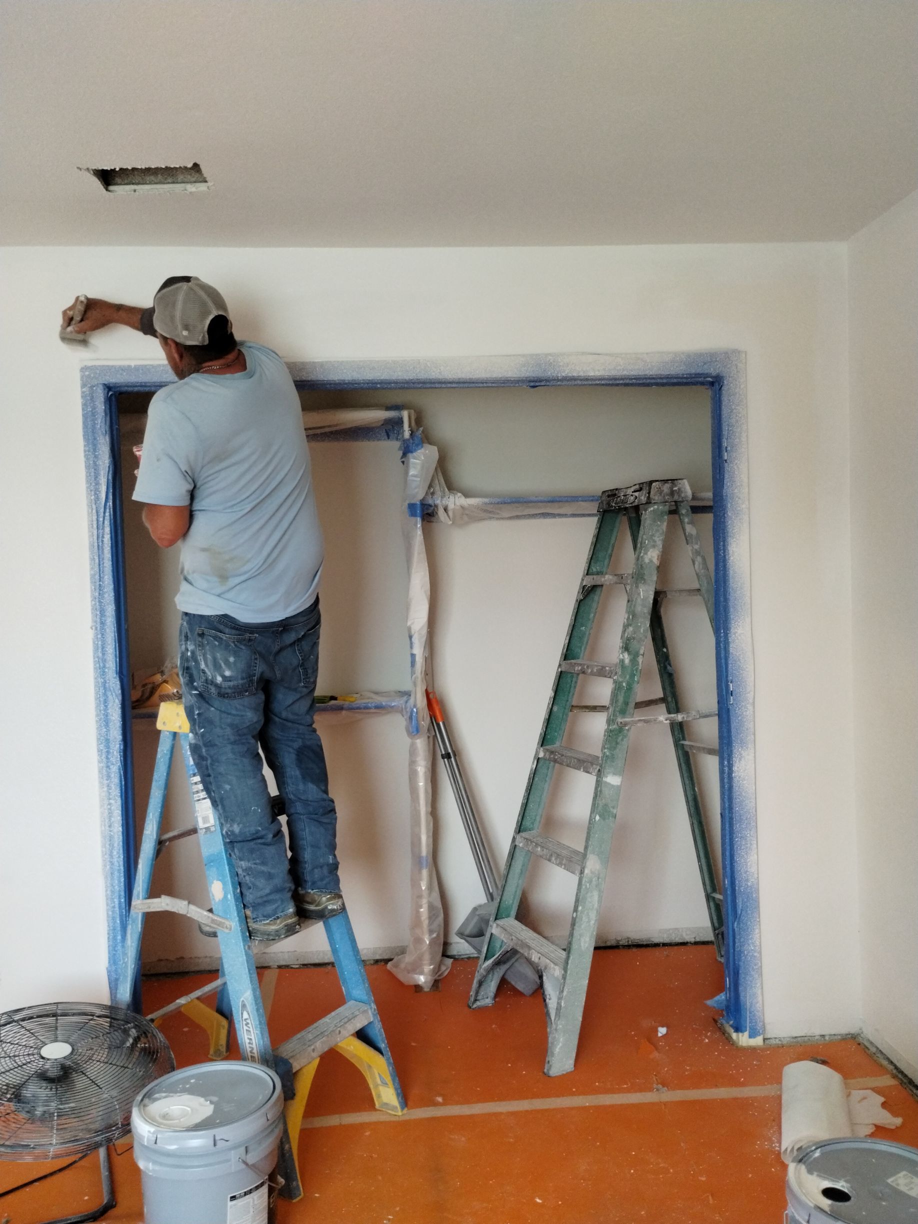 A man painting a room, standing on a blue ladder, near a closet.