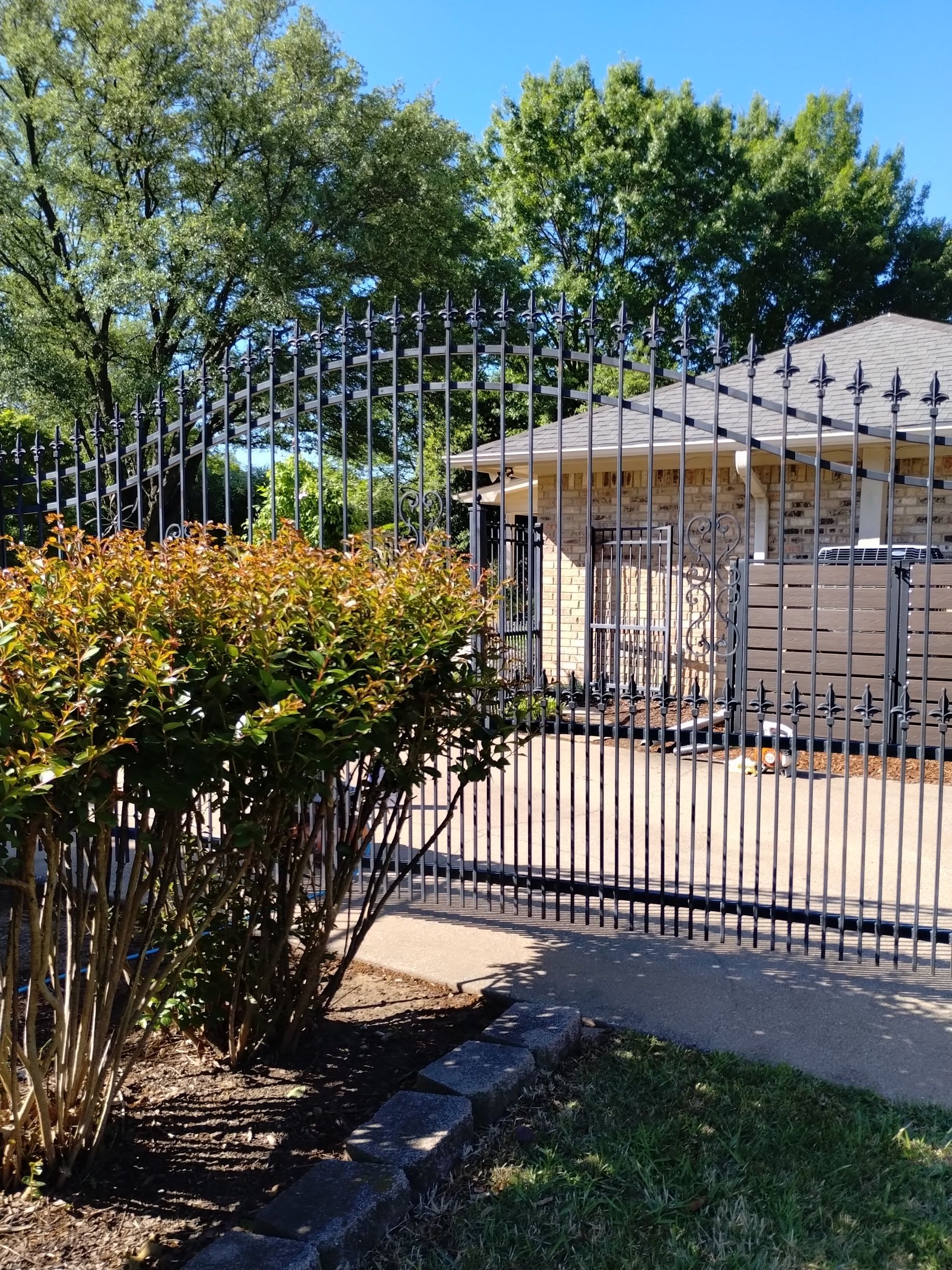 Black metal fence surrounds a brick house, with a bush and trees in the foreground on a sunny day.