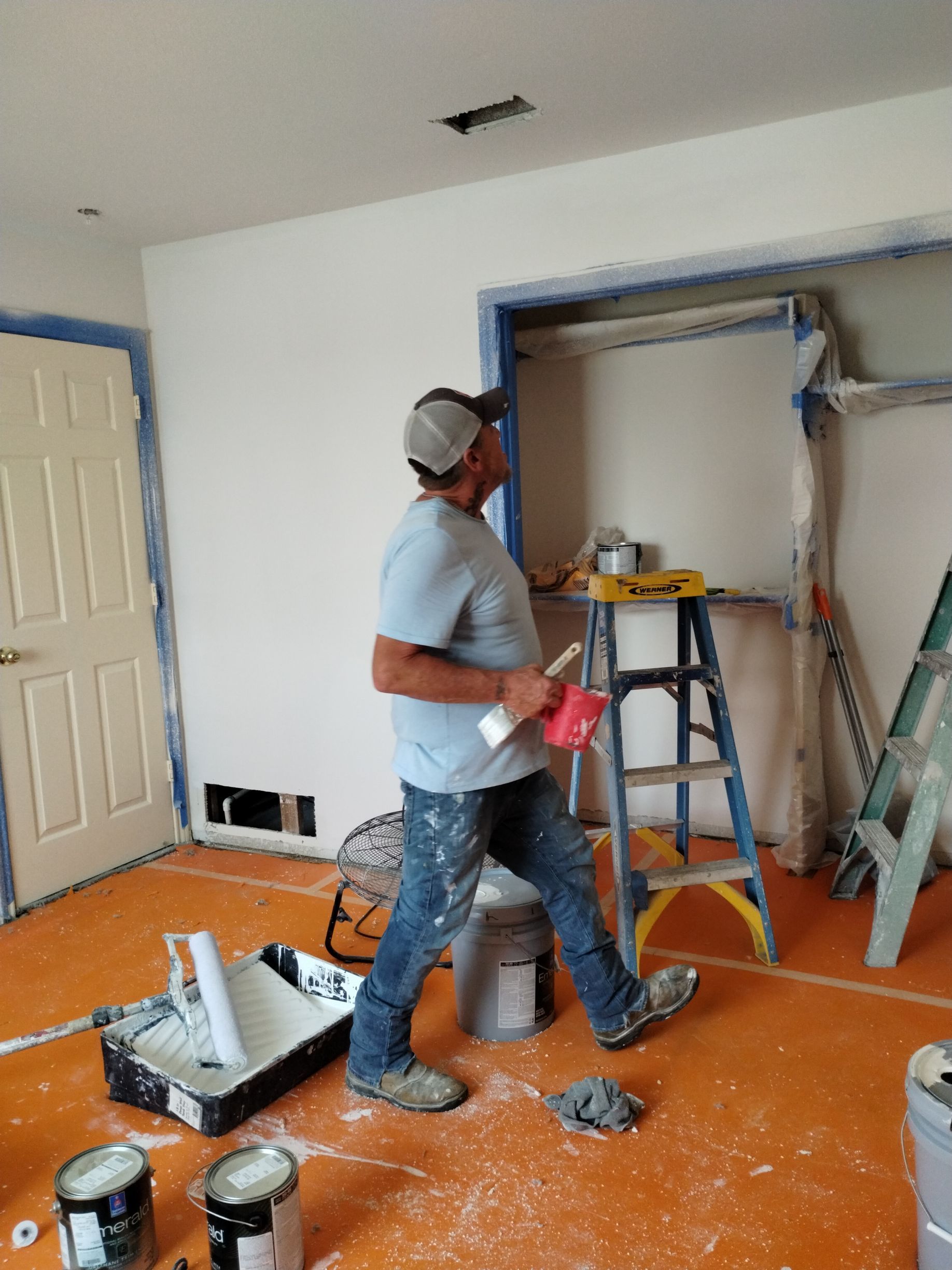 Man in room with paint, tape, and ladders; orange floor, blue trim, white walls.