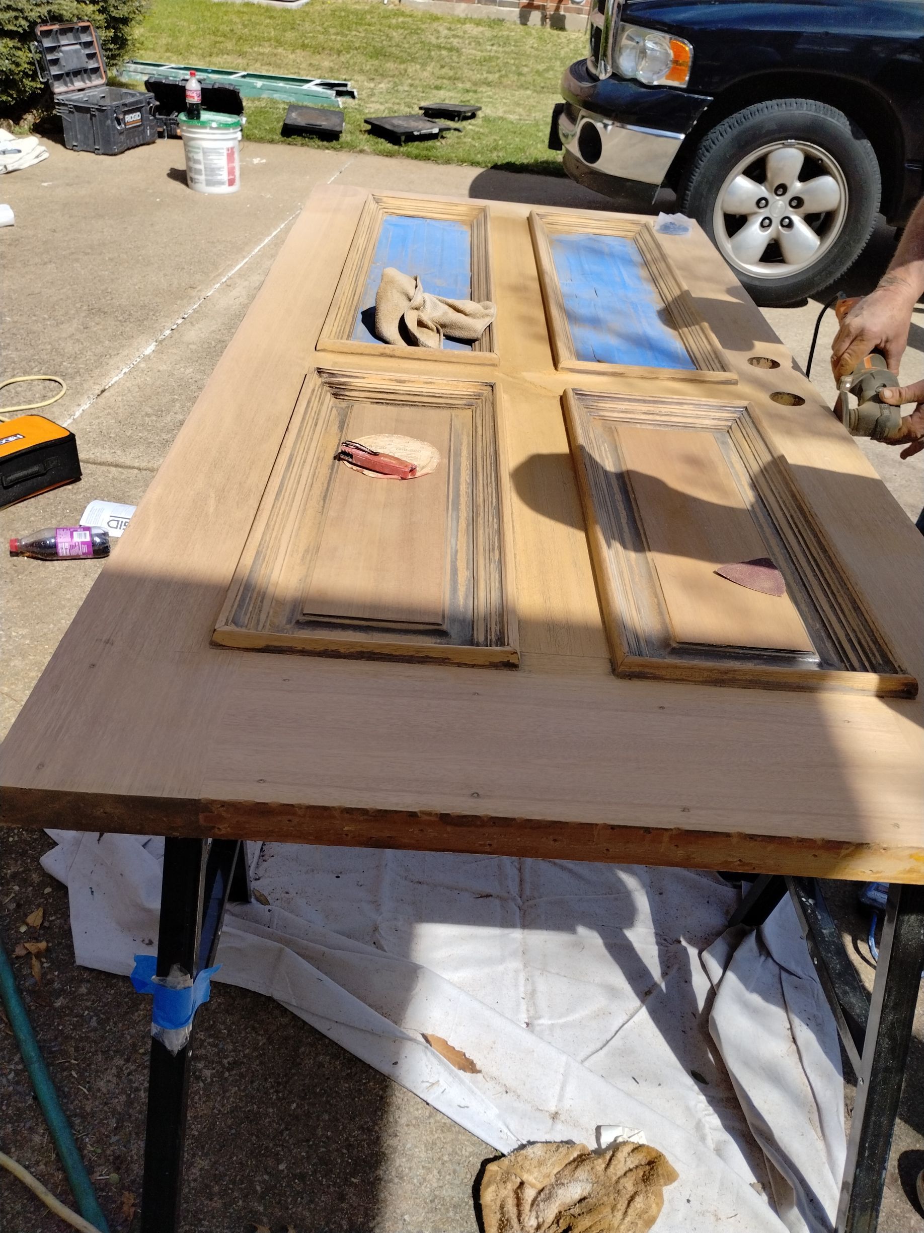 A wooden door on a worktable being sanded, with blue tape over the glass sections, outdoors.
