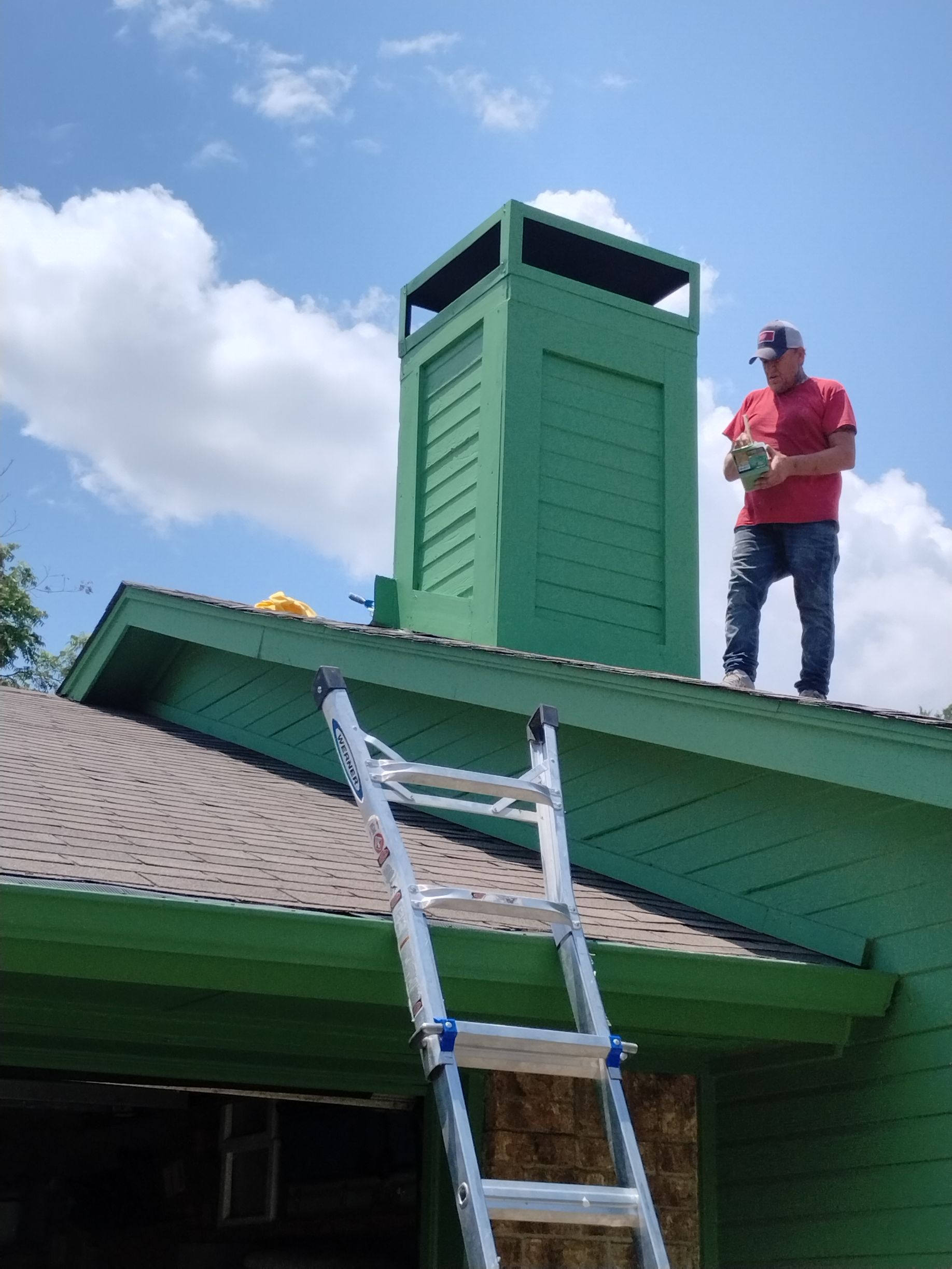 Man on a roof by a green chimney, holding a box, with a ladder on the roof. Bright sky.
