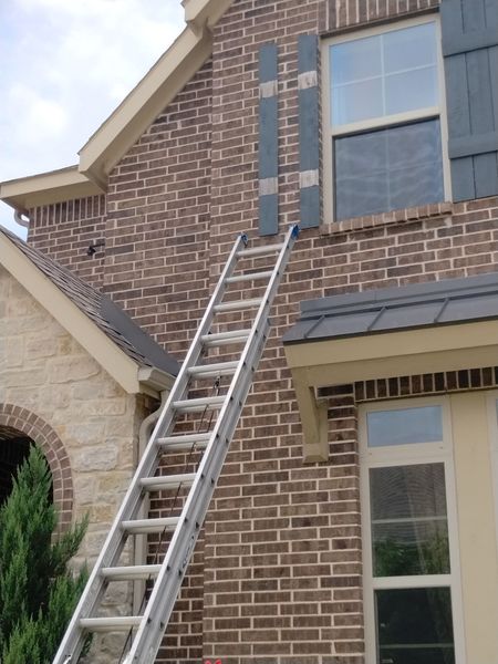 Ladder against a brick house, shutters near window, with a tan roof.
