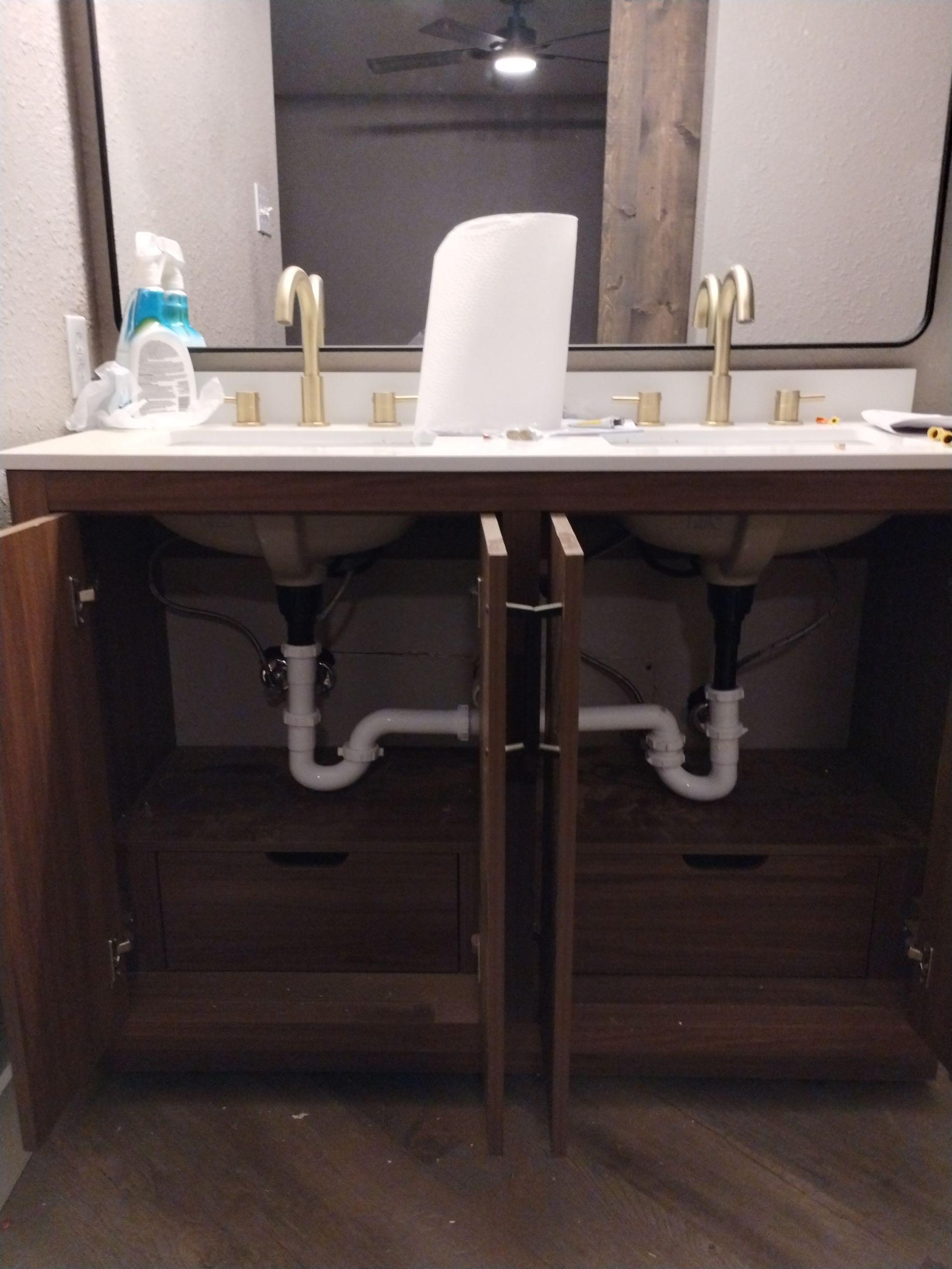 Open cabinet with two sinks, gold faucets, white countertops. Plumbing visible below.