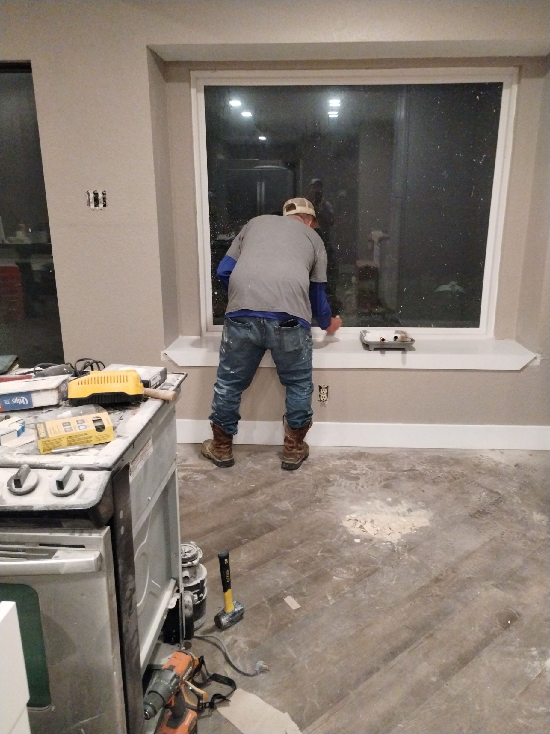 A worker leaning toward a large window inside a room, with construction tools nearby and flooring in progress.