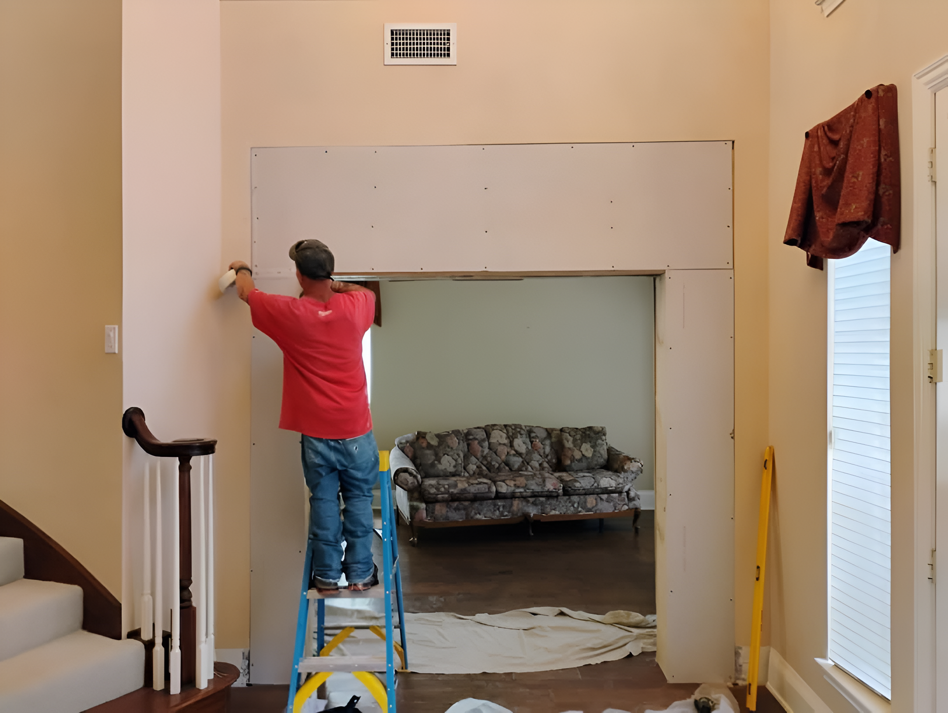 Man on ladder installing drywall in doorway; beige walls, sofa in background.