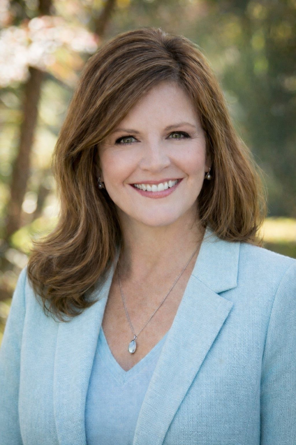 Headshot of a person with shoulder-length brown hair, wearing a light blue blazer, smiling warmly in an outdoor setting.