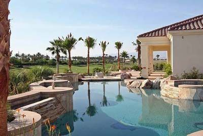 Pool with clear water and reflections, palm trees, and a house with beige walls under a blue sky.