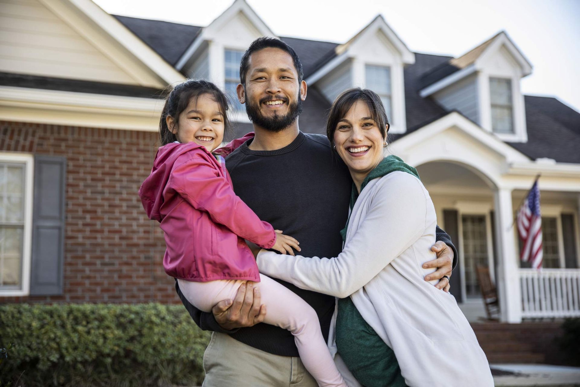 Smiling family stands outside brick home with American flag.