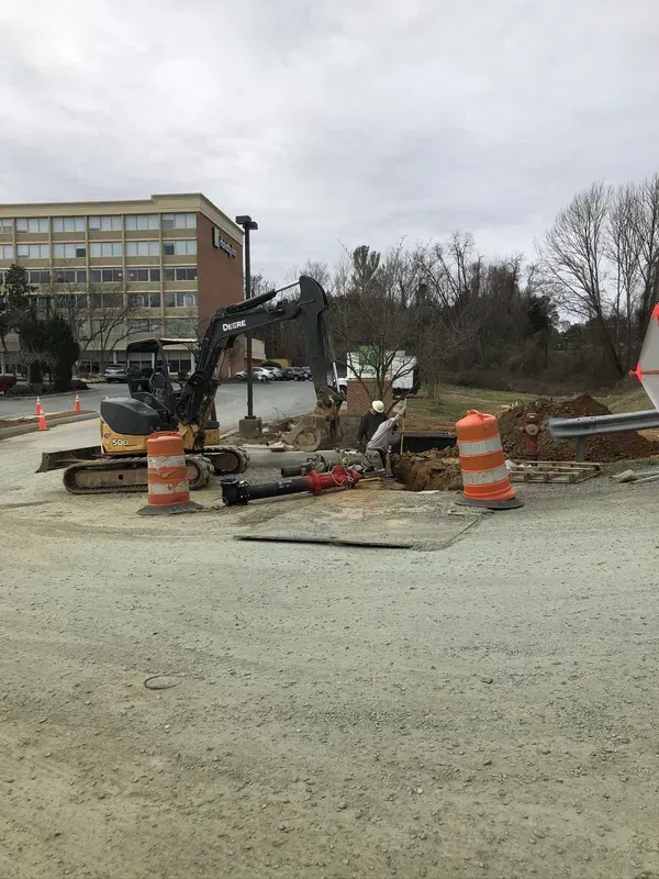 Construction site with excavator digging. Orange traffic cones and a building are visible in the background.
