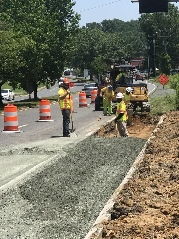 Road construction: Workers in safety vests place gravel along a sidewalk next to a road with orange barrels.