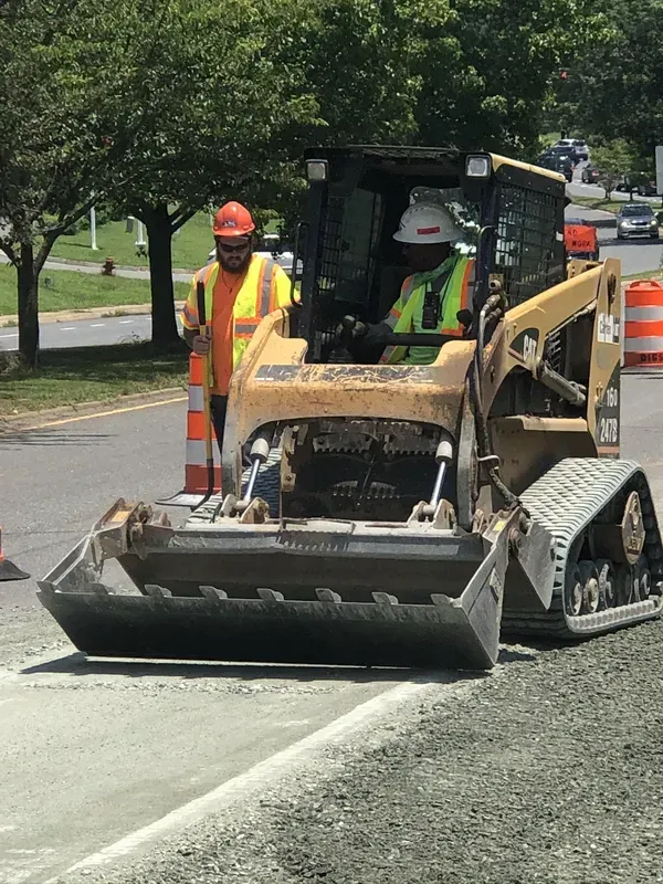 Construction workers operating a skid steer on a road. One worker stands, the other drives.