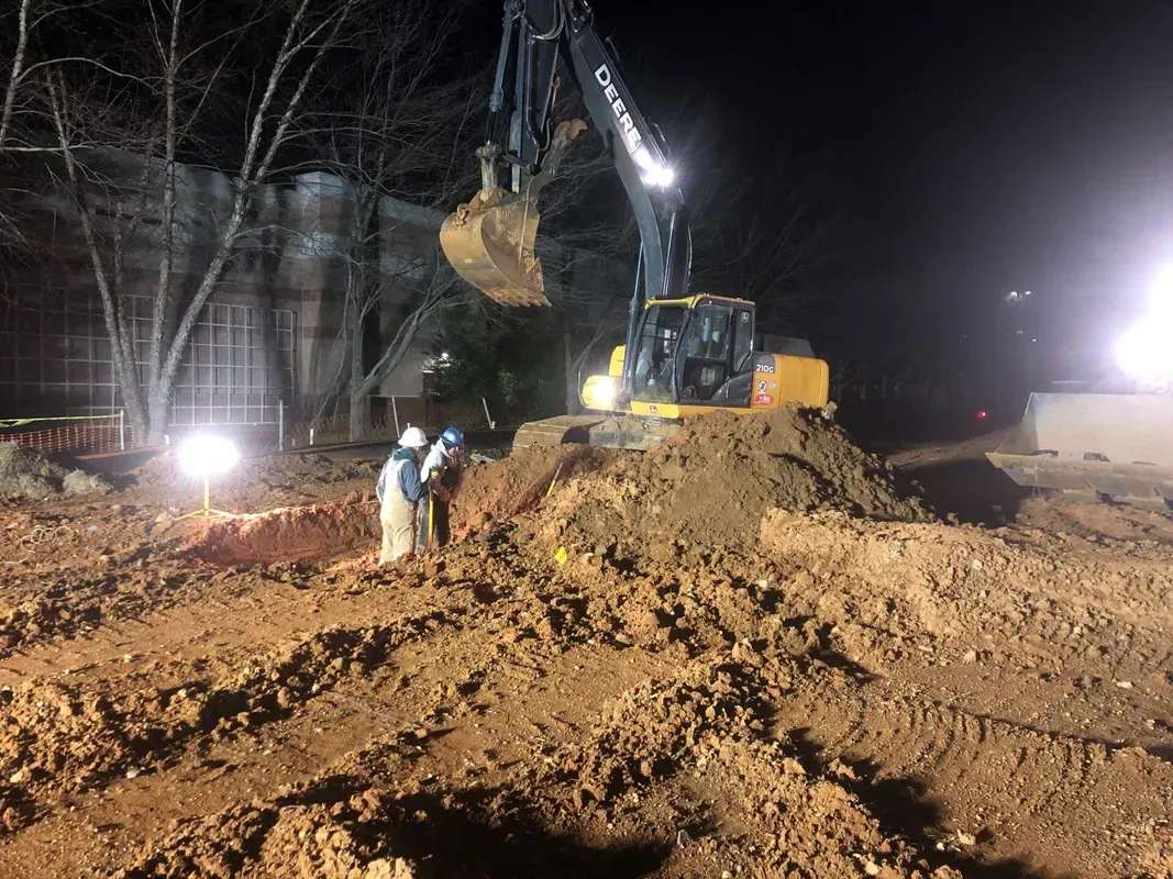 Excavator and workers digging at night with floodlights illuminating the scene. Soil is piled up.