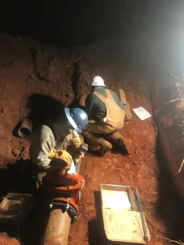 Two workers in a dirt trench, examining plumbing. Dark setting, wearing hard hats and coveralls.