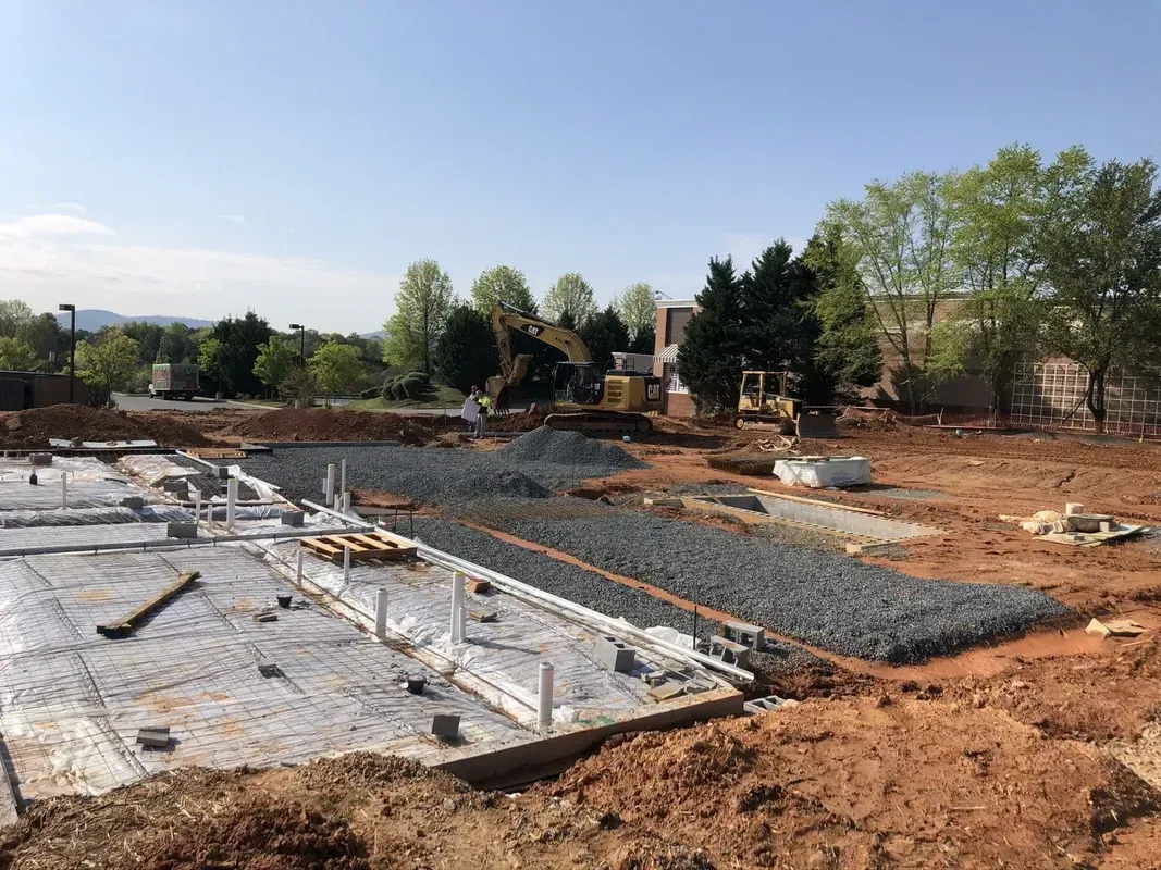 Construction site with foundation work. Gravel, rebar, and pipes visible. Dirt surrounds the foundation.