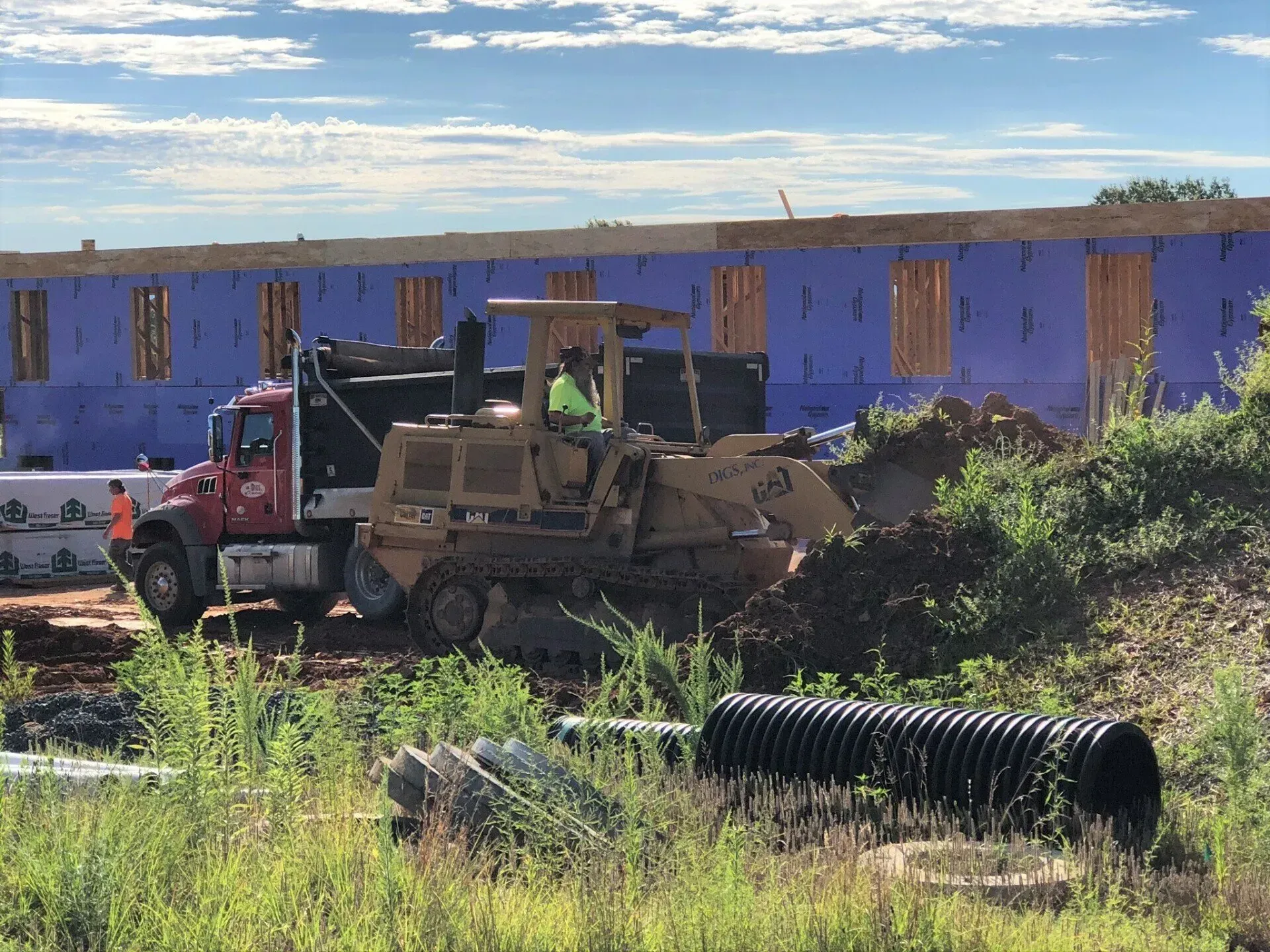 Construction site: Bulldozer loading dirt into a dump truck, building frame in the background, blue sky.