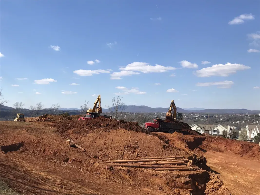 Construction site: Two excavators on a red dirt hill, under a blue sky, with city in the background.