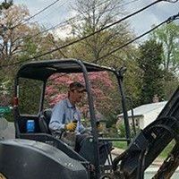 Man operating a small excavator near power lines, trees, and houses.