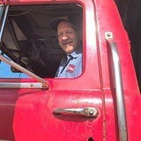 Man in a blue shirt smiles from the driver's seat of a red truck.
