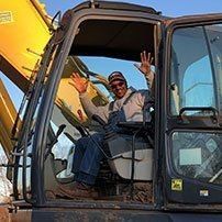 Man waving from excavator cab; sunny day.