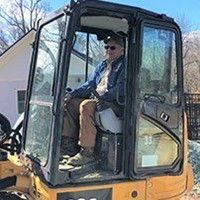 Man operating an excavator in front of a building. He's wearing a blue shirt and sitting inside.