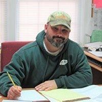 Man in green hoodie and cap, writing at a desk indoors.