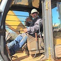 Man in hard hat operating heavy machinery, smiling inside the cab.